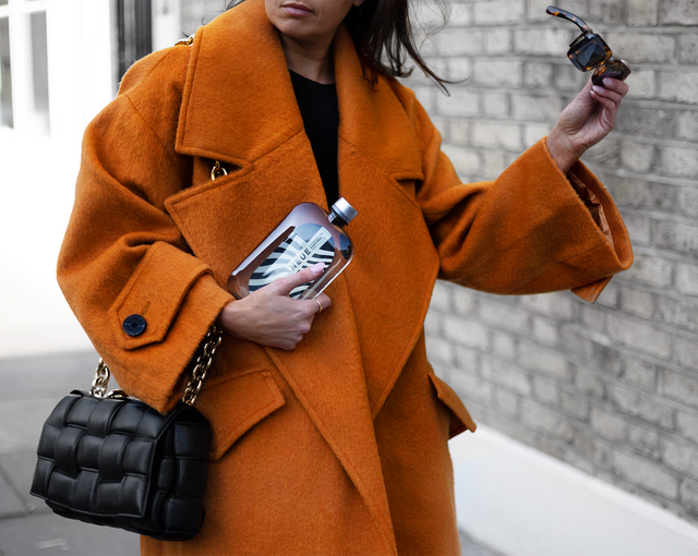 Woman in Orange Coat. Sunglasses. Street Fashion. Woman in a coat wearing a Bottega Venetta Pleated Cassette Bag, sunglasses. Model off duty outfir inspiration